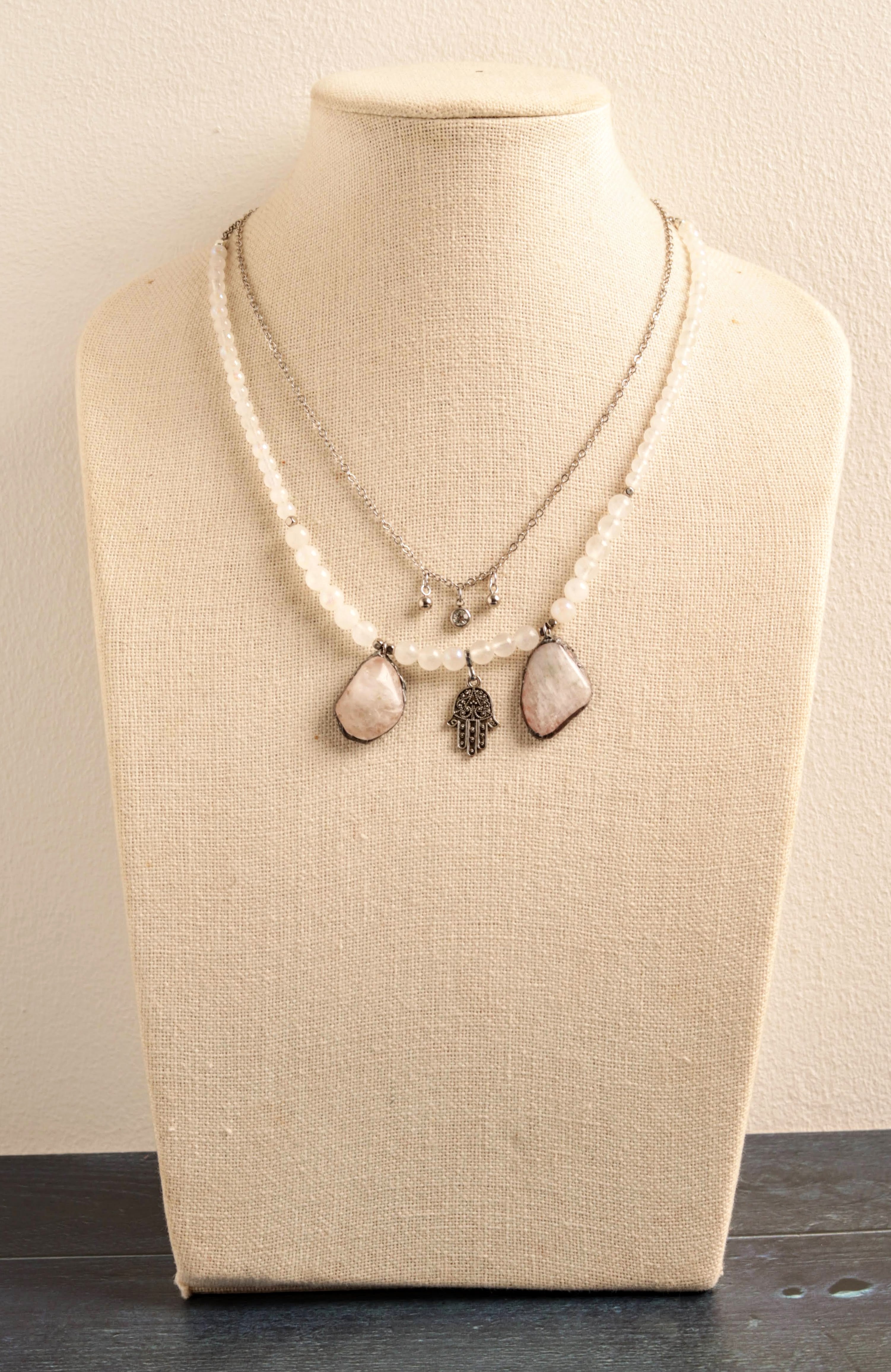 A layered necklace displayed on a cream fabric jewelry bust. The necklace features a double-strand silver-tone chain with a long strand of cream beads, a small silver charm, and two teardrop-shaped cream stone pendants, one with a dark feather charm.