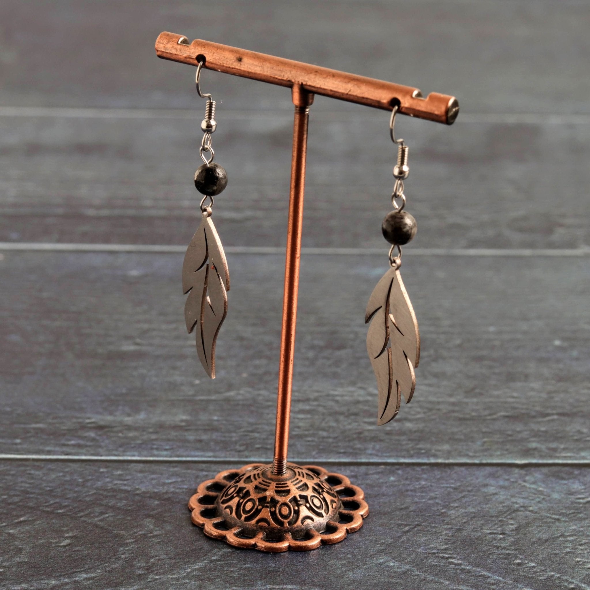 A pair of silver-toned leaf dangle earrings with dark brown beads, displayed on a copper-colored metal stand against a dark gray wood plank background.